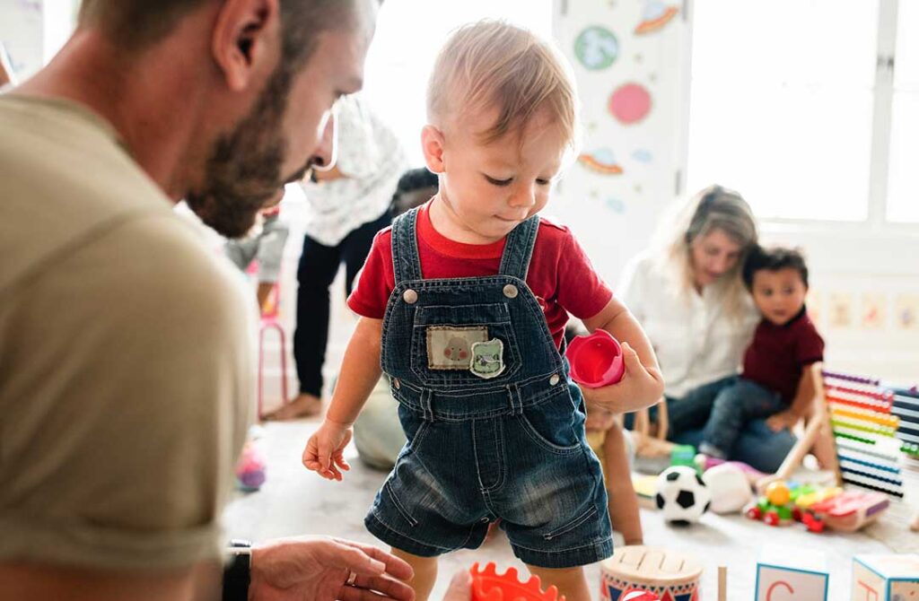 Preschool children build a colorful block structure while an educator observes and asks questions.